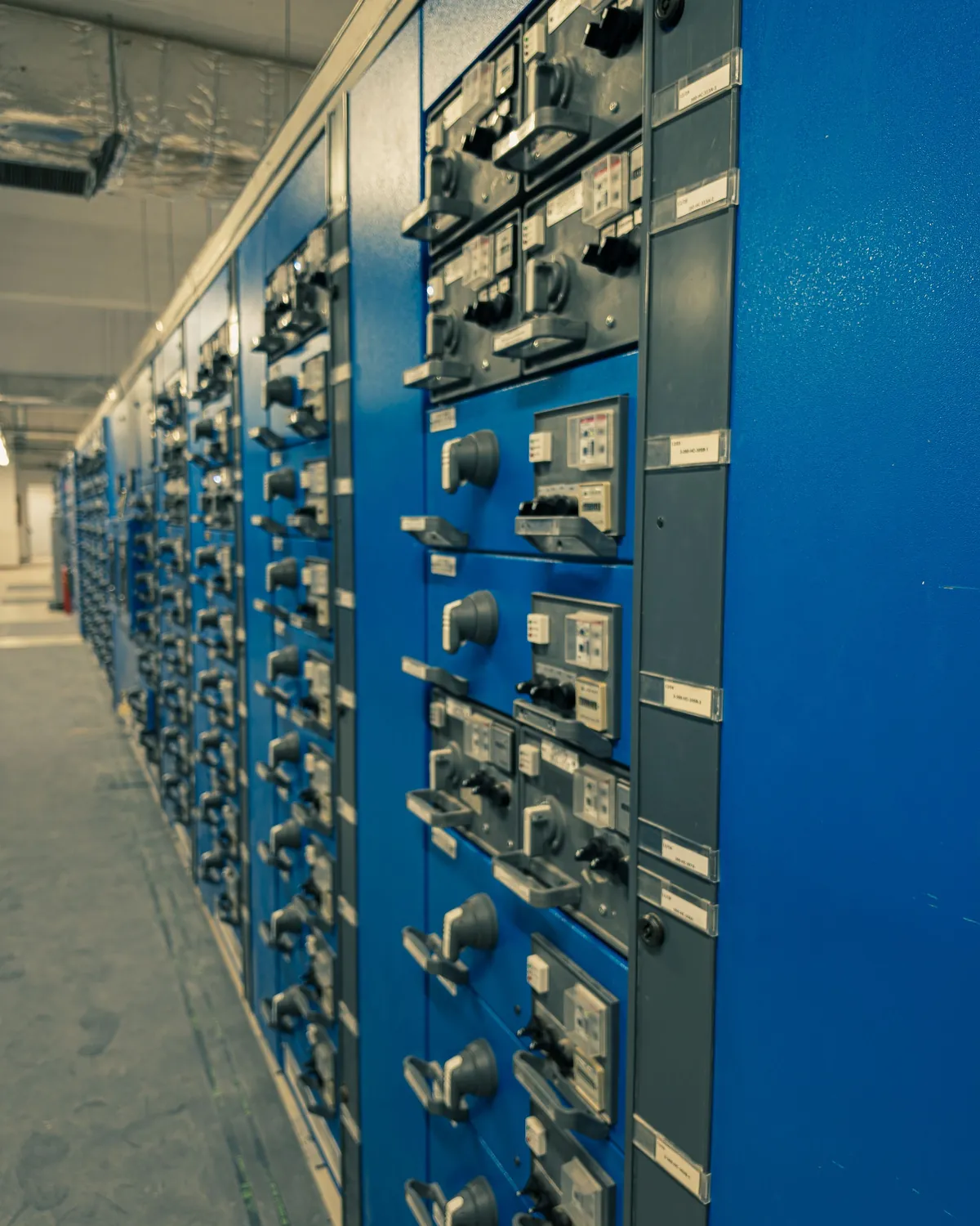 Rząd niebieskich szaf rozdzielnicy elektrycznej z wyłącznikami i opisanymi polami w pomieszczeniu rozdzielni.