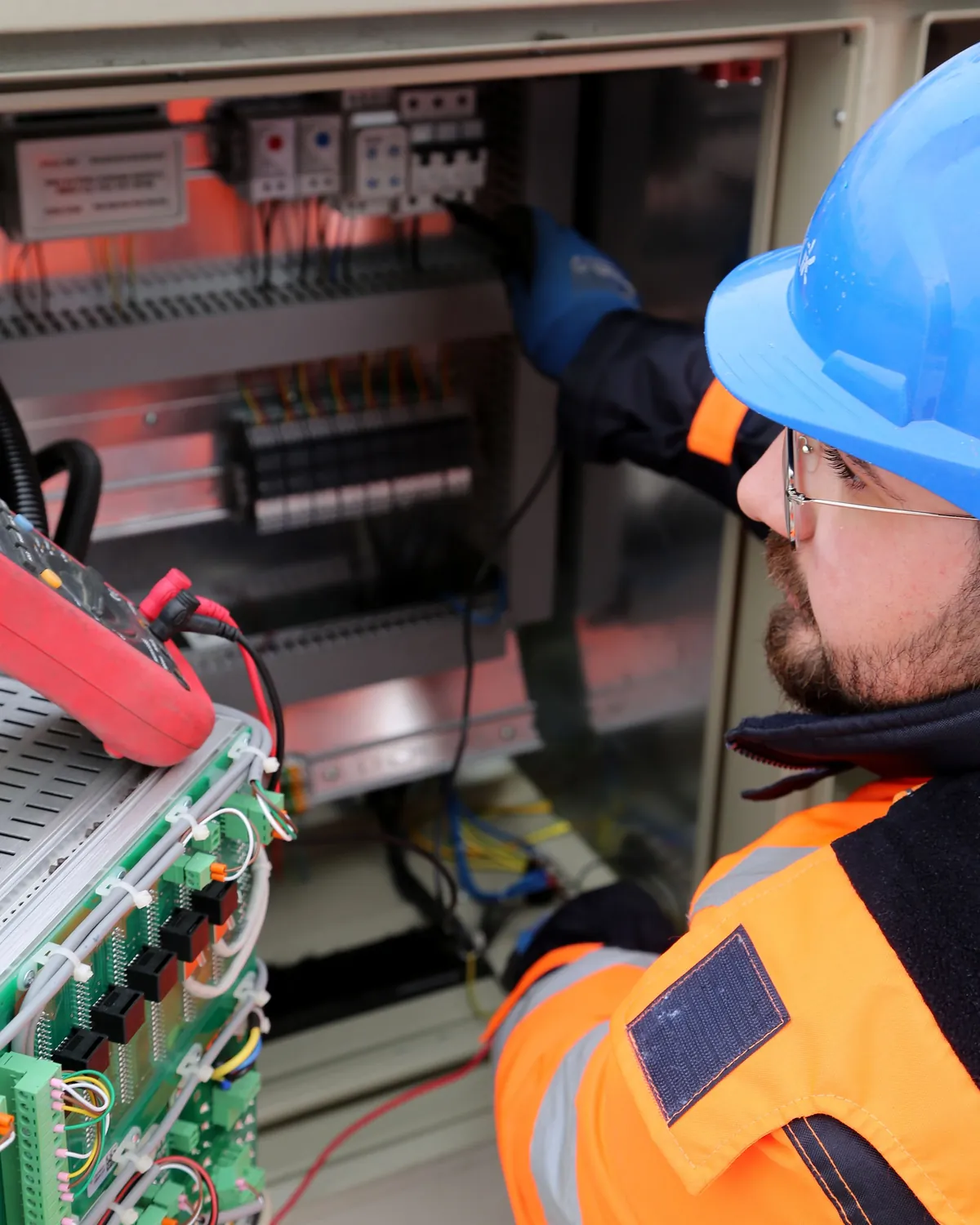 Elektryk w kasku, okularach ochronnych, rękawicach i odzieży ostrzegawczej wykonuje pomiar w otwartej szafie sterowniczej.